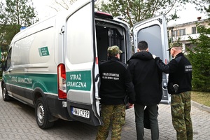 Zatrzymany obywatel Egiptu Dwóch funkcjonariuszy Straży Granicznej oraz jeden cudzoziemiec stoją tyłem, przed nimi pojazd służbowy Straży Granicznej.