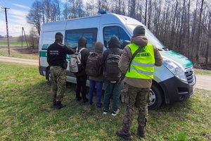 Zatrzymani cudzoziemcy Zatrzymani cudzoziemcy, funkcjonariusz Straży Granicznej, żołnierz WOT, a za nimi auto służbowe Straży Granicznej.