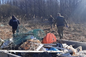 Akcja sprzątania rzeki Biebrzy Dwóch funkcjonariuszy Straży Granicznej oraz jeden Straży Pożarnej poruszają się po zarośniętym terenie podczas akcji porządkowej. Na pierwszym planie widać kontener wypełniony rożnymi odpadami.