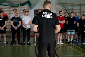 I Ty możesz zostać funkcjonariuszem Straży Granicznej Na zdjęciu w sali gimnastycznej stoi funkcjonariusz Straży Granicznej przodem do grupy mężczyzn w czarnym stroju sportowym. Przed nim ustawiona jest grupa młodych mężczyzn w sportowych ubraniach, którzy słuchają go w skupieniu. W tle widać drabinki gimnastyczne i elementy wyposażenia.